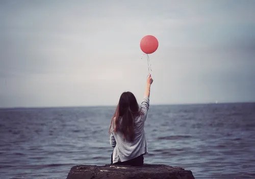 A woman by the sea releasing a red balloon as a symbol of healing, self-forgiveness and personal growth.